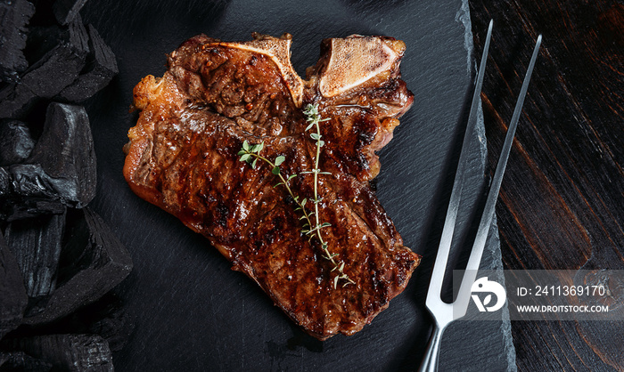 barbeque steak on a black slate board with meat fork and grill coals next to it