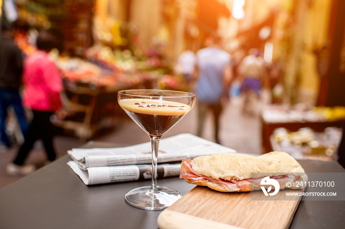 Shakerato drink in cocktail glass with panini and newspaper on the table. Italian lunch with traditional coffee and sandwich with prosciutto