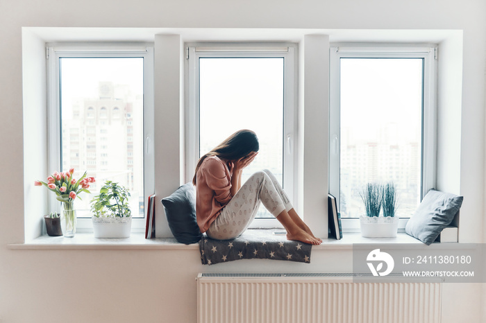 Sad young woman crying about something while sitting on the window sill at home