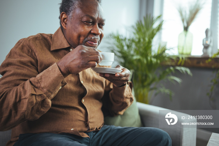 Thoughtful older man sitting on couch at home alone, holding cup of tea or coffee, feeling lonely and sad, unhappy depressed mature male looking into distance, thinking about problems