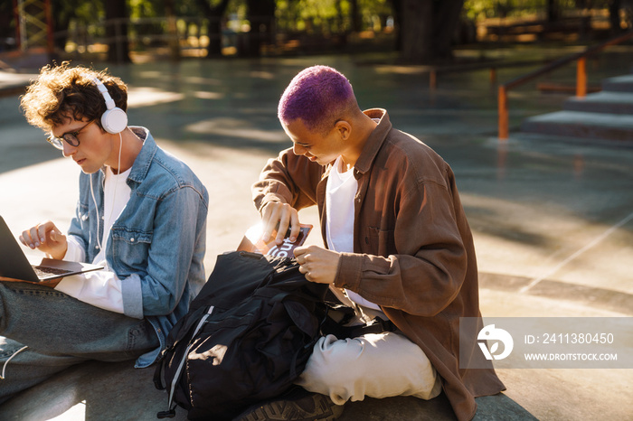 Two young handsome stylish students sitting in skatepark