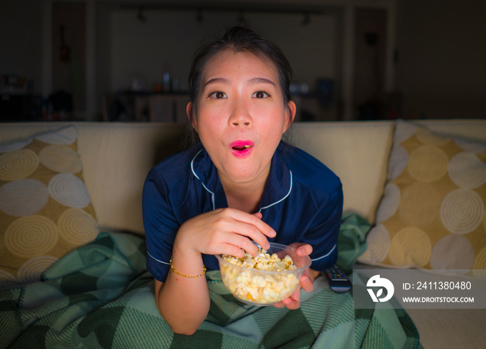 young beautiful happy and relaxed Asian Japanese woman at home living room sitting cozy on sofa couch watching TV show episode or romantic comedy movie eating popcorn