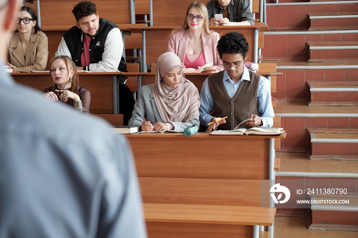 Several rows of contemporary intercultural university students sitting by long wooden desks in lecture hall and listening to professor at lesson