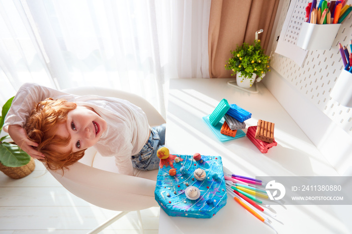 happy kid sitting at the desk with handcrafted solar system, made of modeling plasticine