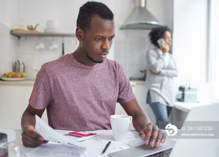 Young dark-skinned male managing family budget at home, paying gas and electricity bills online for the first time, looking at laptop screen, while his wife calling to bank. Family budget and debts