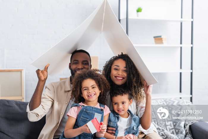 happy african american family looking at camera under paper roof in new apartment