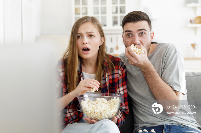 Young guy and girl looking at TV screen holding popcorn bowl and eating, showing great amazement, fear and disappointment, being very attentive to what is going on