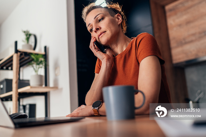 Worried and sad woman sitting at the kitchen table and talking on the phone