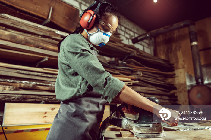 Busy and serious craftswoman grinding timbers with special machine. Beautiful woman wearing safety glasses. Concept of joiner’s shop and woodworking. Gender equality. Male profession