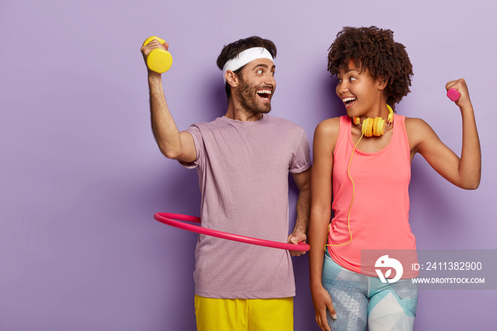 Horizontal shot of happy mixed race woman and man have exercises indoor, train biceps, dressed in active wear, guy rotates hula hoop, lead healthy lifestyle, attend gym regulary, isolated on purple