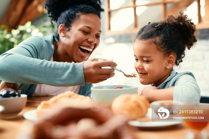 Cheerful African American mother feeds her small daughter during breakfast at home.