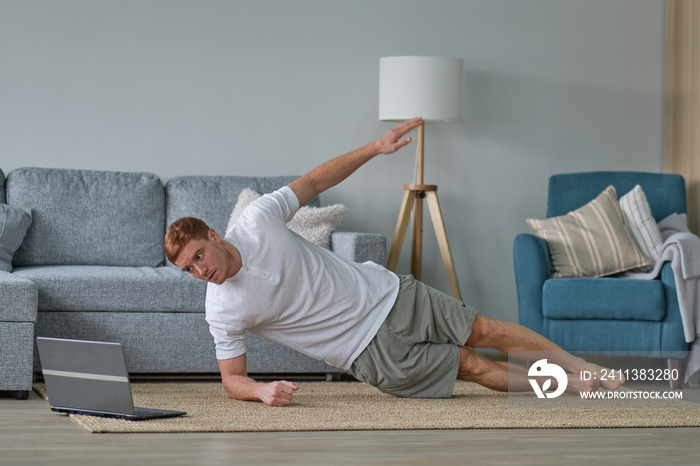 young active man watching online video training on laptop, exercising, stretching during morning workout at home. Sport, healthy lifestyle. Get Fit At Home.