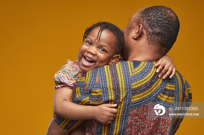 African happy little girl smiling at camera while embracing her dad on yellow background