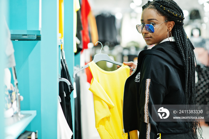 Afican american women in tracksuits and sunglasses shopping at sportswear mall against shelves. She choose yellow t-shirt. Sport store theme.