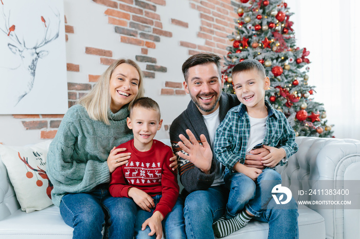 young family of four men woman husband and two little boys sons happily having fun together looking at webcam video communication with friends sitting on sofa at home near christmas and new year tree