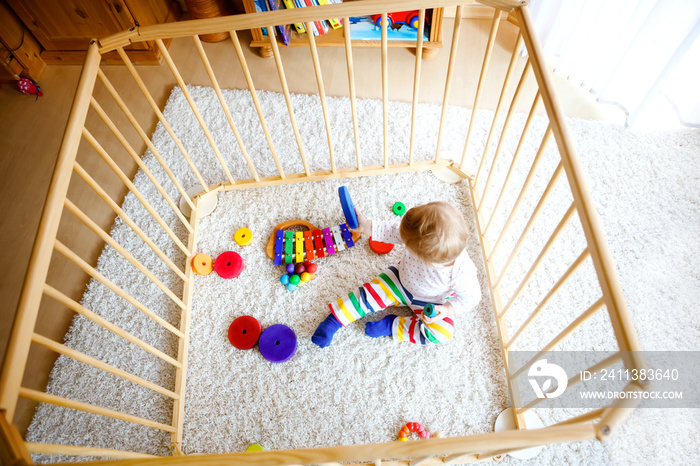 Beautiful little baby girl standing inside playpen. Cute adorable child playing with colorful toys. Home or nursery, safety for kids. Alone baby waiting for mom