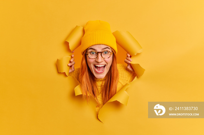 Excited cheerful redhead millennial woman stares surprisingly at camera has overjoyed expression keeps mouth opened wears trendy bright hat transparent glasses poses through torn paper background