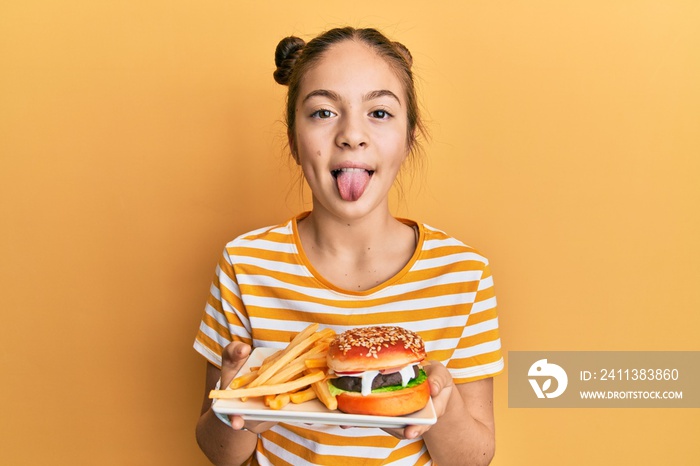 Beautiful brunette little girl eating a tasty classic burger with fries sticking tongue out happy with funny expression.