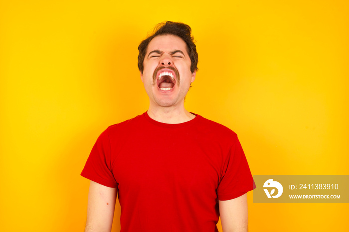 Young Caucasian man wearing red t-shirt standing against yellow wall angry and mad screaming frustrated and furious, shouting with anger. Rage and aggressive concept.