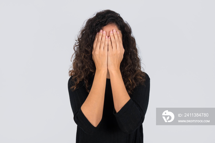 Curly haired brunette woman standing dressed in black  covering her face with her hands over gray background.