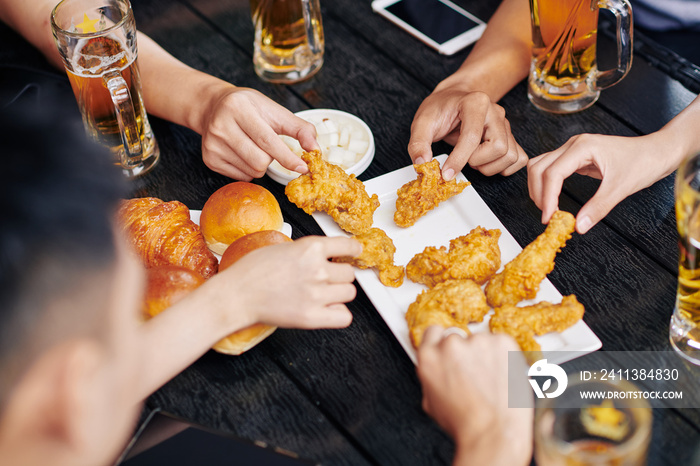 Close-up of people sitting at the wooden table drinking beer and eating fried chicken together in cafe