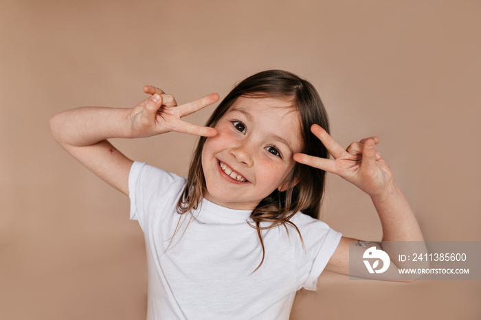 Portrait of pretty charming girl showing peace signs near the face and smiling over beige background. Happy true emotions, isolated background