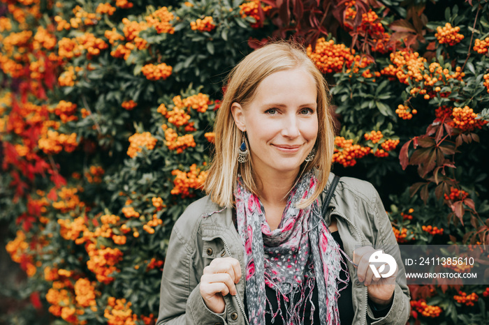 Outdoor portrait of beautiful blond woman, posing next to bright orange scarlet firethorn berries (Pyracantha coccinea), wearing khaki color parka and pink scarf