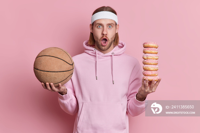 Surprised man in sweatshirt holds basketball and pile of sweet high calories doughnuts chooses between sport and unhealthy food keeps mmouth opened isolated over pink background. hobby leisure