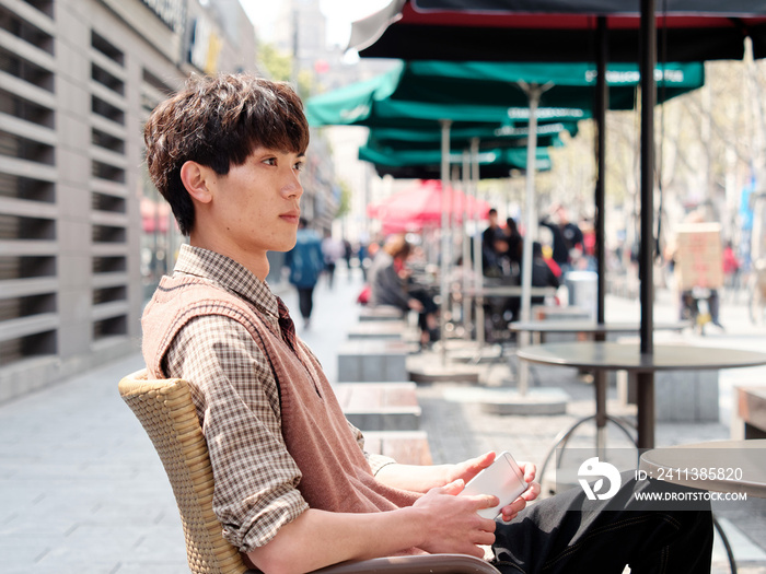 Portrait of handsome Chinese young man with curly black hair in plaid shirt and wool vest sitting outdoor cafe with his mobile phone in hand in sunny day, male fashion, cool Asian young man lifestyle