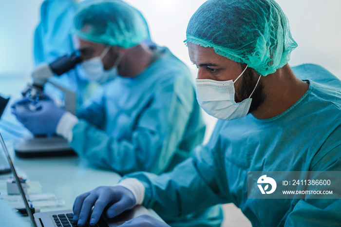 Medical workers in hazmat suit working with laptop computer and micoroscope inside laboratory hospital during coronavirus outbreak - Focus on right man face