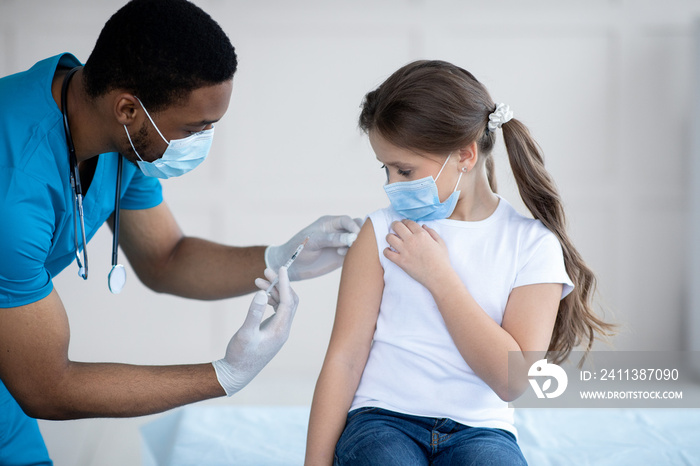 Young black doctor making covid-19 vaccine injection to little girl in face mask at hospital