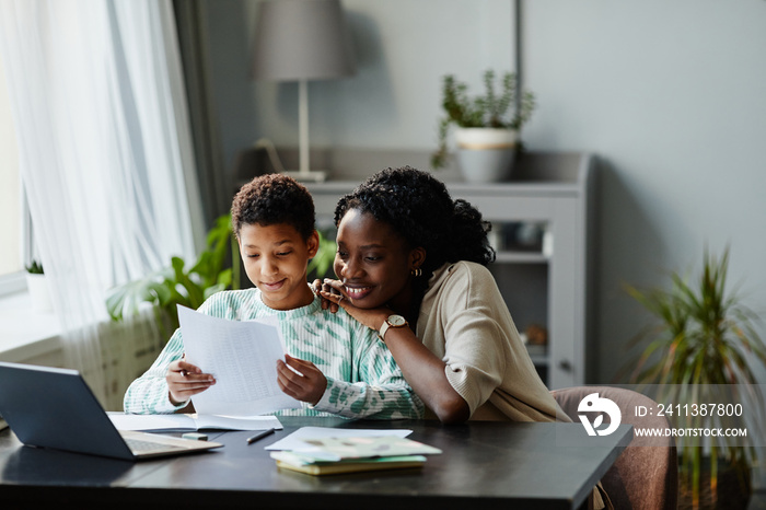 Front view portrait of smiling teenage girl holding test result while studying at home with mother