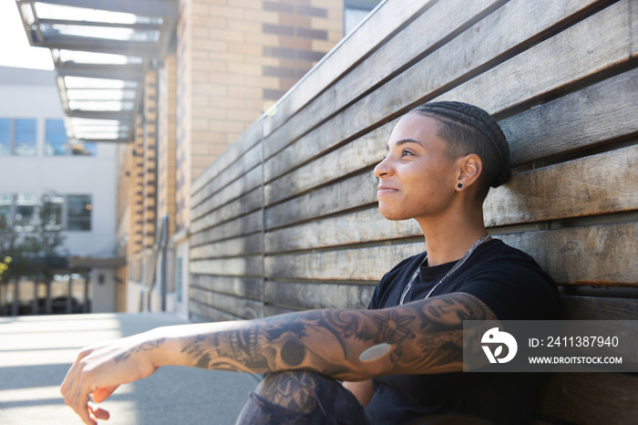 Young African American woman with tattoos on arm wearing black tee shirt sitting against wood wall outside