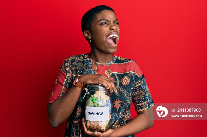 Young african american woman holding savings jar with south african rands money angry and mad screaming frustrated and furious, shouting with anger. rage and aggressive concept.