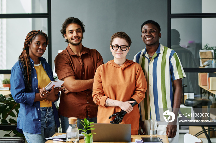 Group of young creative designers in casualwear standing by workplace with laptop and supplies in office and looking at camera