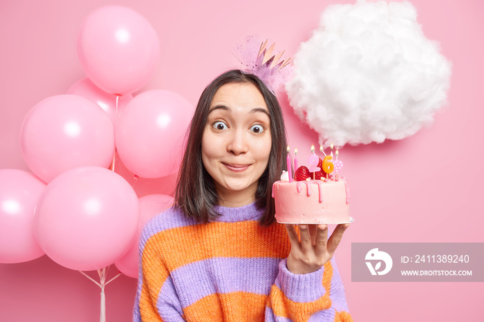 Surprised brunette birthday girl holds delicious cake with burning candles didnt expect to receive so many presents wears casual jumper and crown on head poses near balloons being on festive party