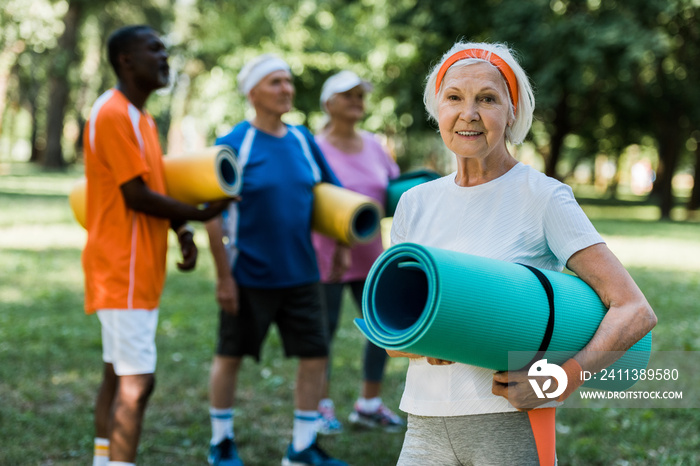 selective focus of happy retired woman with fitness mat smiling near multicultural pensioners