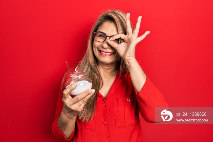 Middle age hispanic woman holding bowl with sugar smiling happy doing ok sign with hand on eye looking through fingers