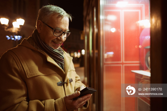 Senior man is looking at store front and typing on phone.