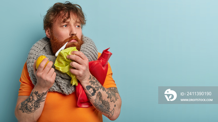 Indoor shot of thoughtful puzzled man measures temperature with digital thermomemeter in mouth, holds hot water bottle, tissue, wears knitted scarf, caught cold and fever, suffers from headache