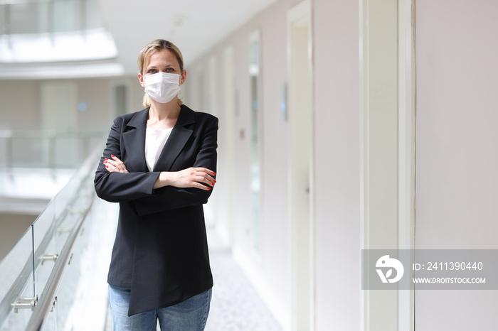 Businesswoman in medical protective mask stands with folded arms in hotel corridor