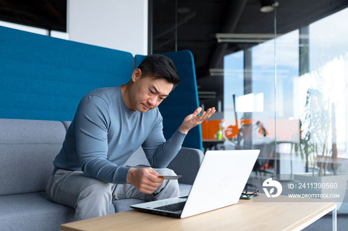 Annoyed and frustrated Asian man trying to make an online purchase sitting in the office at the computer, using a credit card