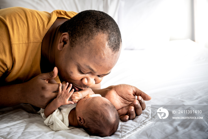 An African American father kissing hand, his 12-day-old baby newborn son lying in bed in a white bedroom, with happy, concept to African American family and newborn