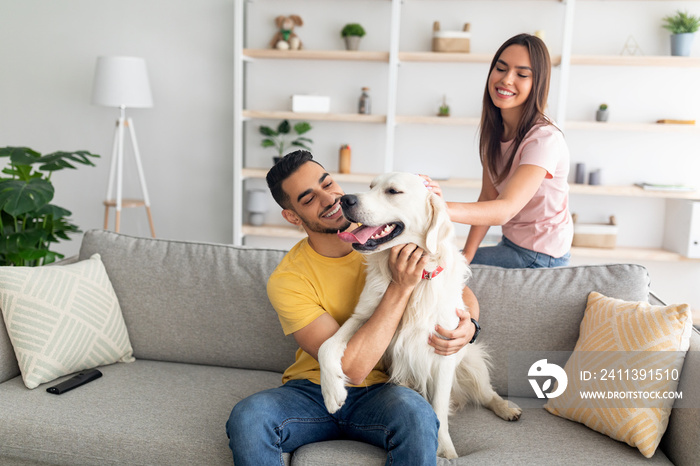 Joyful young diverse couple stroking their cute dog on couch at home, empty space