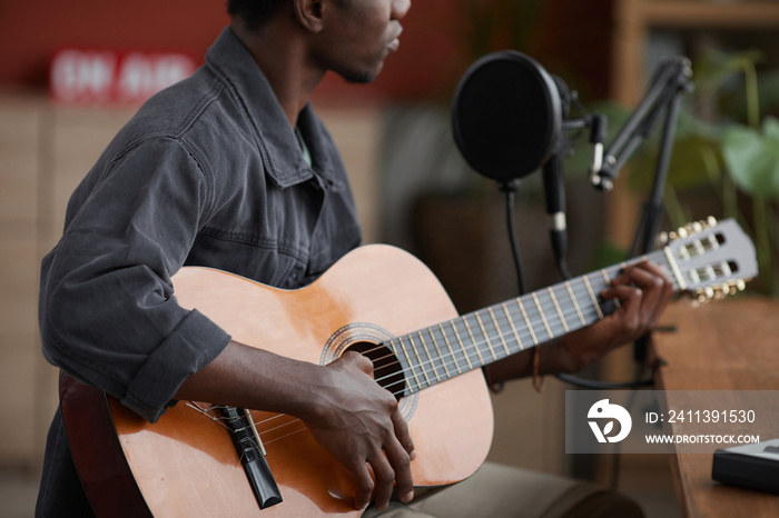 Cropped portrait of young African-American man playing guitar while sitting by microphone in home recording studio, copy space