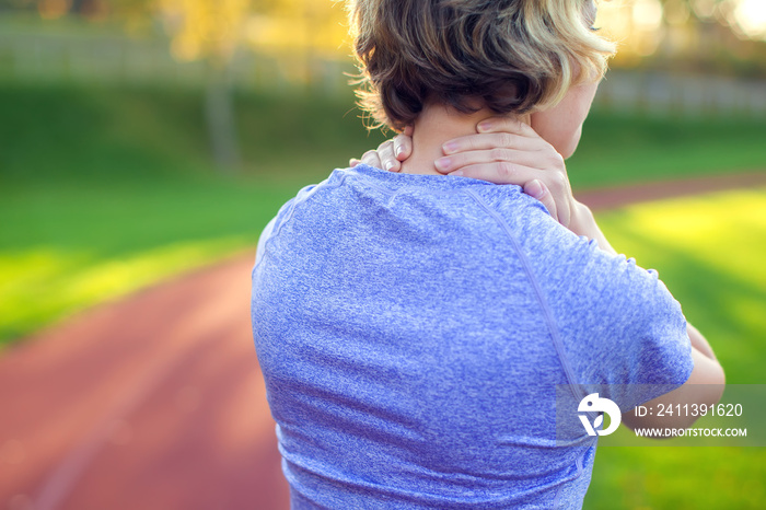 Back view of athletic young woman in sportswear touching her painful neck at the stadium. Sport, health and people concept