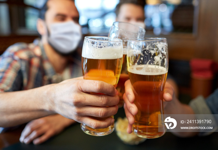 people, leisure and pandemic concept - male friends wearing face protective medical masks for protection from virus disease drinking beer and clinking glasses at bar or pub