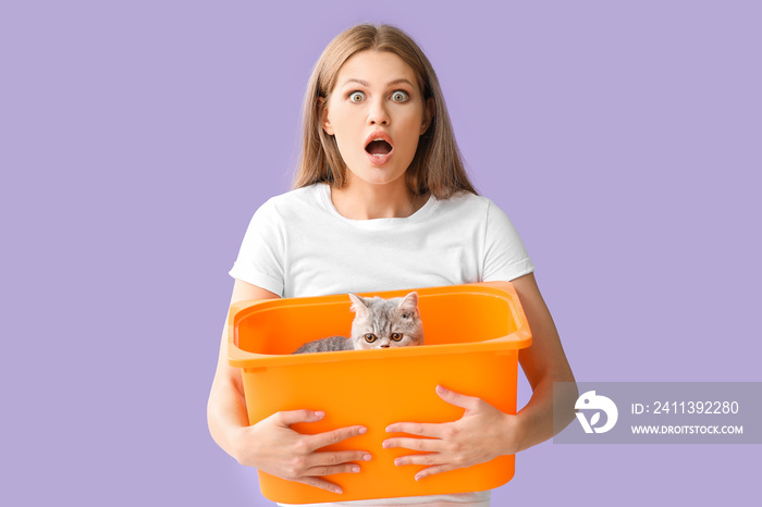 Shocked young woman holding box with cute cat on color background