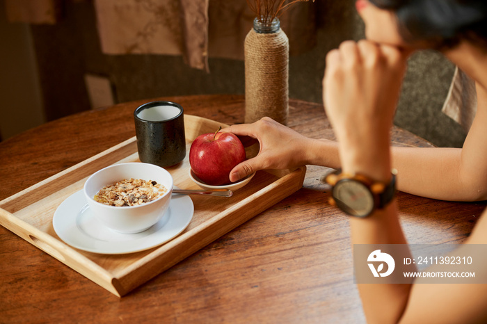 Woman taking red apple from tray with healthy breakfast consisting of porridge and cup of warm water