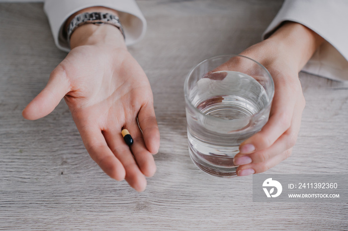 Cropped image of woman’s hands holding pills and glass of water indoor under wooden table. Pharmacy and healthcare. Female holding drugs and pure water.